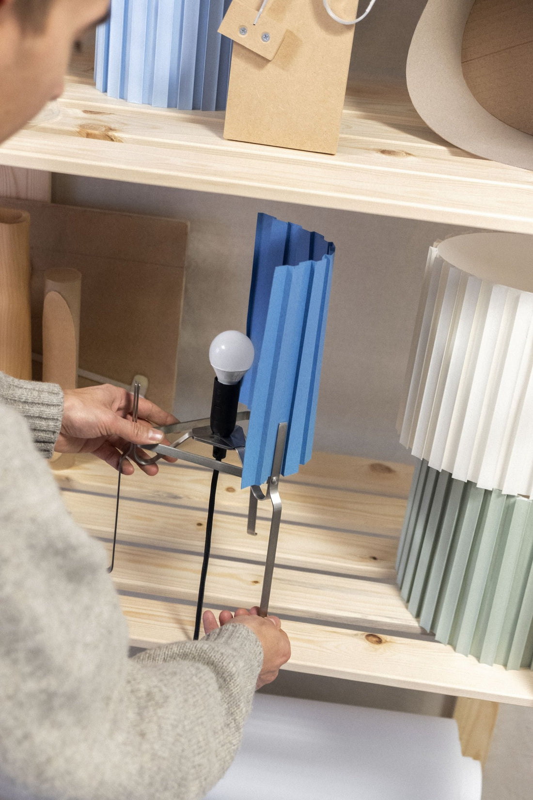 A designer adjusting the frame of a lamp prototype beside pleated shade samples on a wooden shelf.
