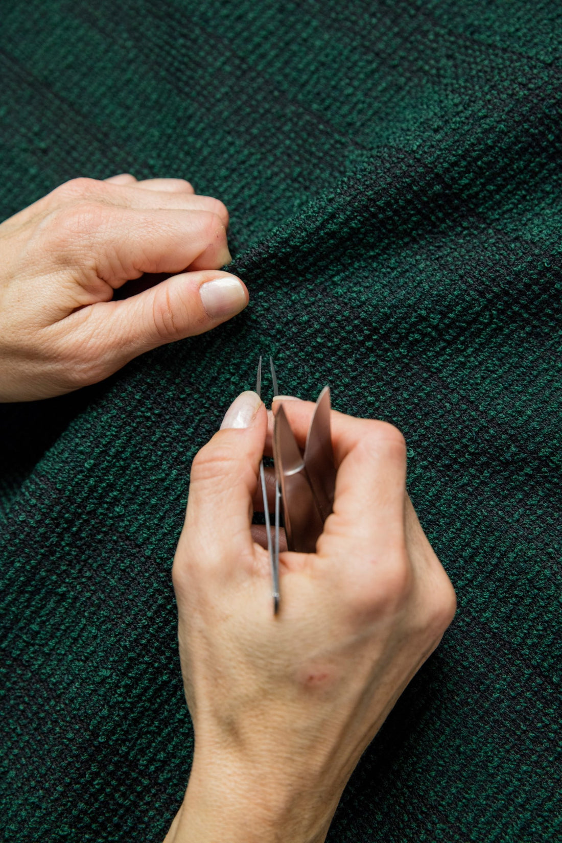 Hands inspect and mend a woven wool fabric with tweezers.