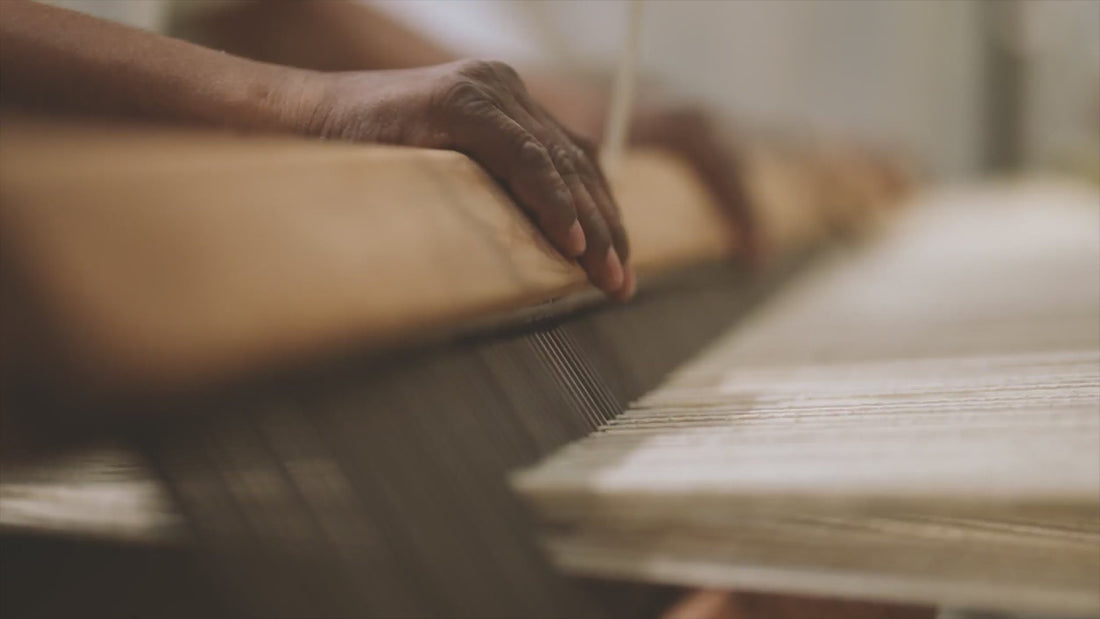 A weaver's hands at a hand loom press the reed against the rug as it is woven.