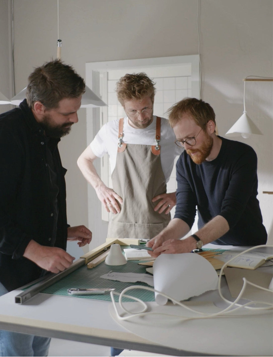 Three designers working on lamp prototypes at a workbench, with pendant shades hanging behind them.