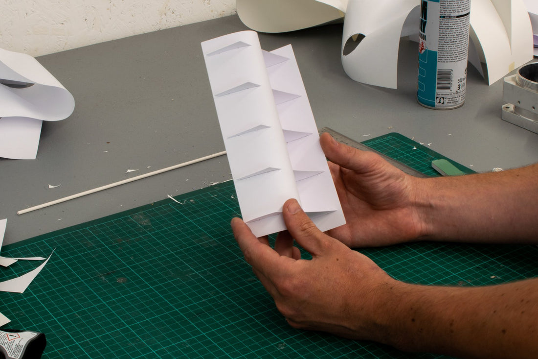 Hands fold a paper model for the Tension series on a cutting mat in the studio, more models behind.