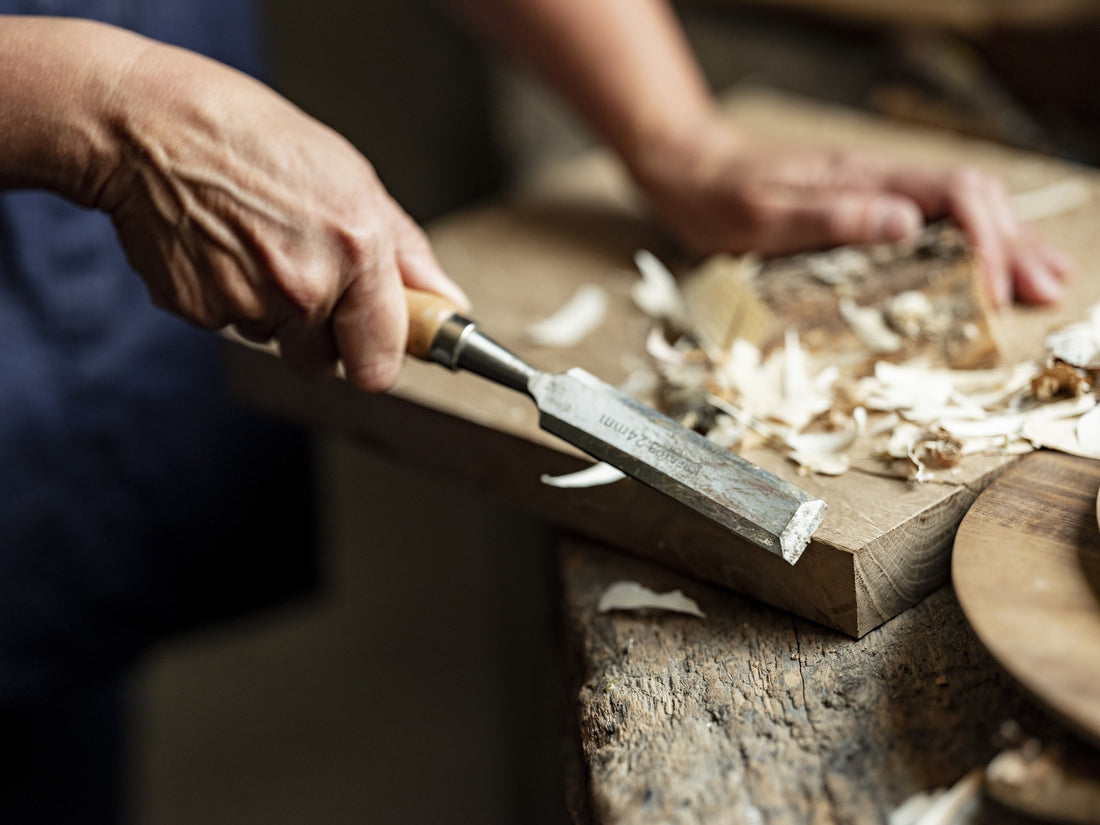 Two hands work a chisel along the edge of a wooden board on a rustic workbench.