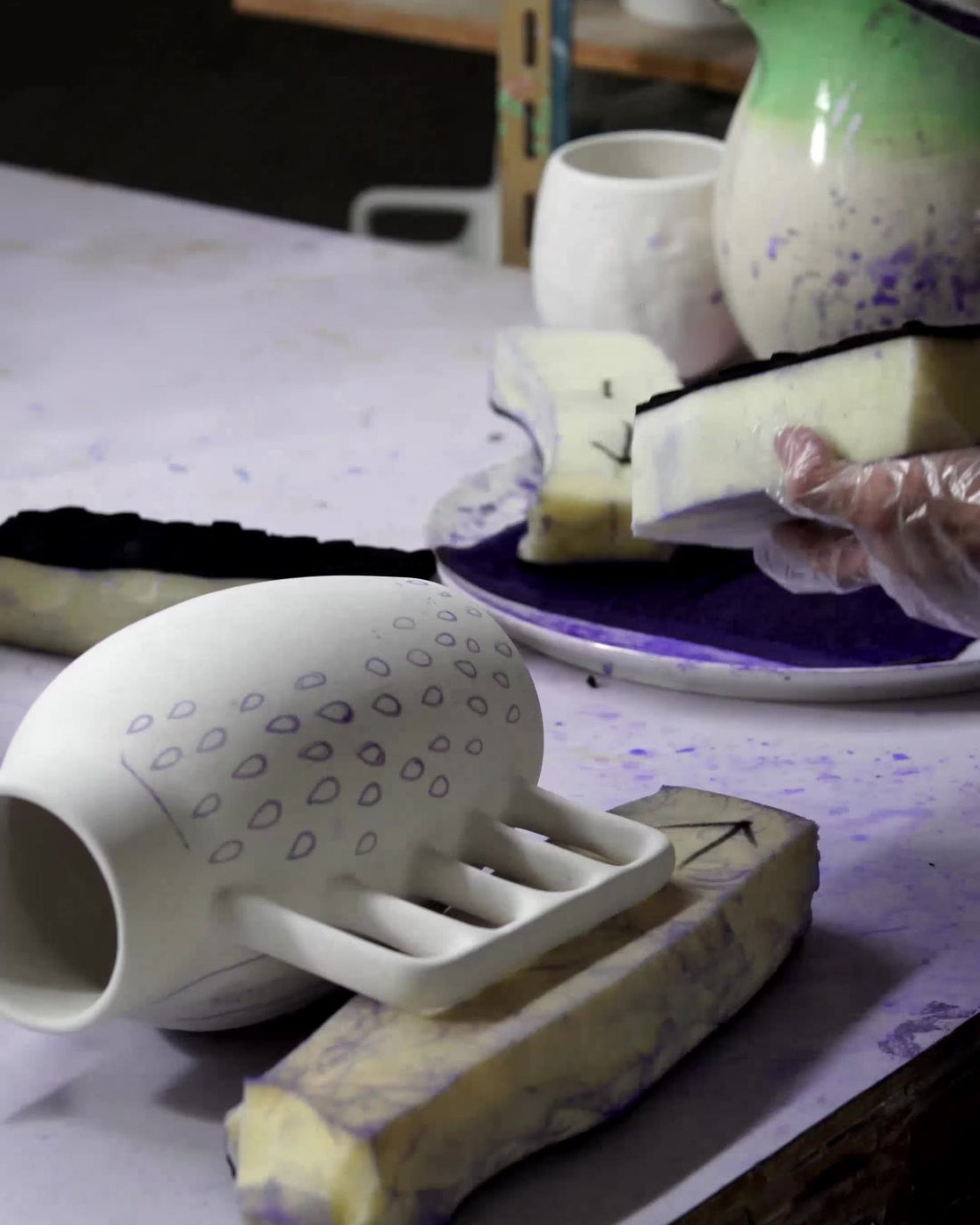 A raw ceramic vase with a drawn pattern on the worktable, glaze being applied with sponge and glove behind.