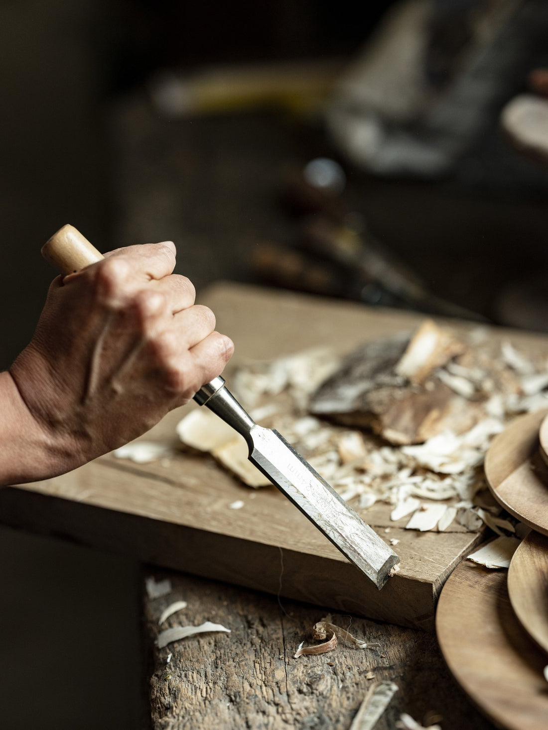 A hand guides a chisel across a wooden board, surrounded by fresh wood shavings.