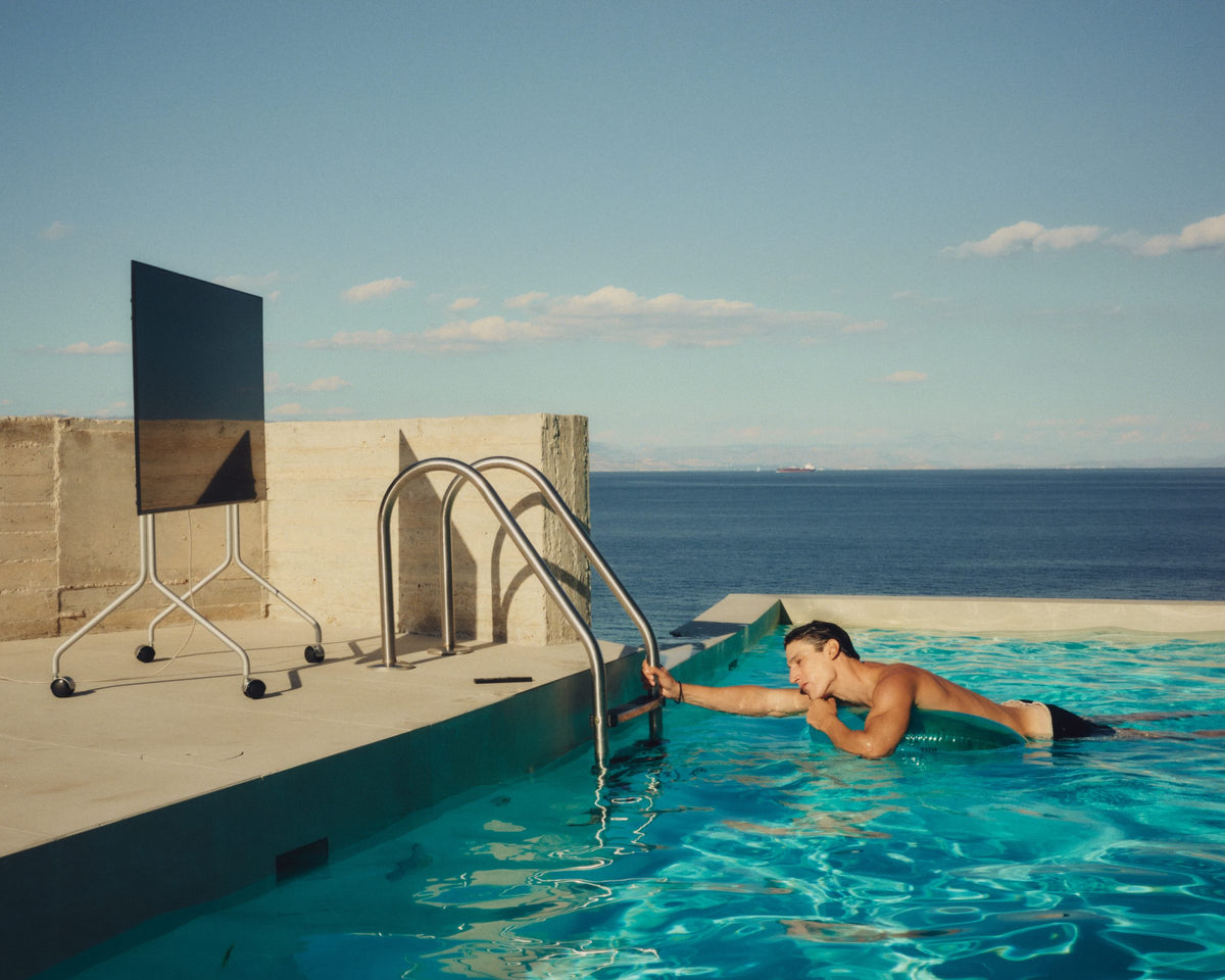 A Pedestal Moon TV stand on a concrete terrace beside a pool by the sea, a man in the water holding the pool ladder, still from Objects of Desire